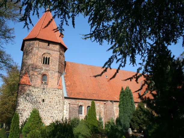 St. Laurentius-Kirche Backsteinkirche mit roten Dachziegeln umgeben von Nadelbäumen bei blauem Himmel. Turm mit Schießscharten.