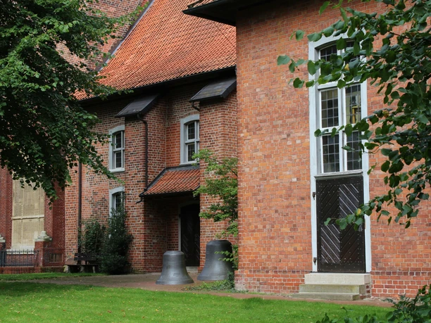 Backsteinkirche mit roten Dachziegeln, umgeben von Grün, Kirchturmglocken am Eingang platziert.