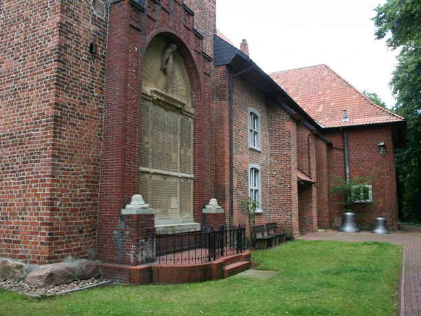 Backsteinkirche mit gotischen Elementen, angrenzendem Kirchgarten und zwei großen Glocken im Freien.