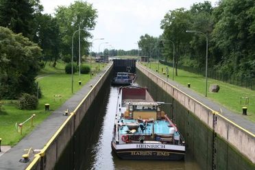 Kanal mit Schleuse und zwei Frachtschiffen unter blauem Himmel, umgeben von Bäumen und Grünflächen.