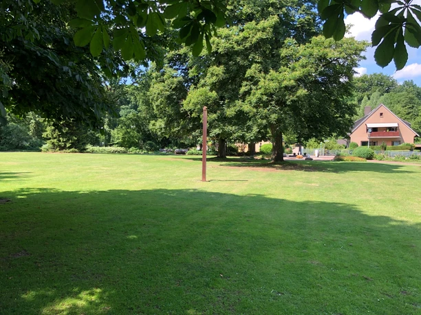 Eine gepflegte Rasenfläche mit Bäumen, im Hintergrund ein Satteldachhaus am Waldrand bei sonnigem Wetter.