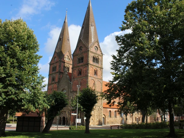 Zweiturmige romanische Kirche aus Backstein mit Spitzdach, umgeben von Bäumen und blauen Himmel.