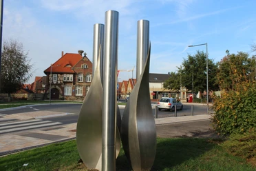 Skulptur Stundenrot schenkt nichts als Blätter Drei hohe, spiralförmige Metallsäulen stehen in einem städtischen Umfeld mit traditionellen Gebäuden.