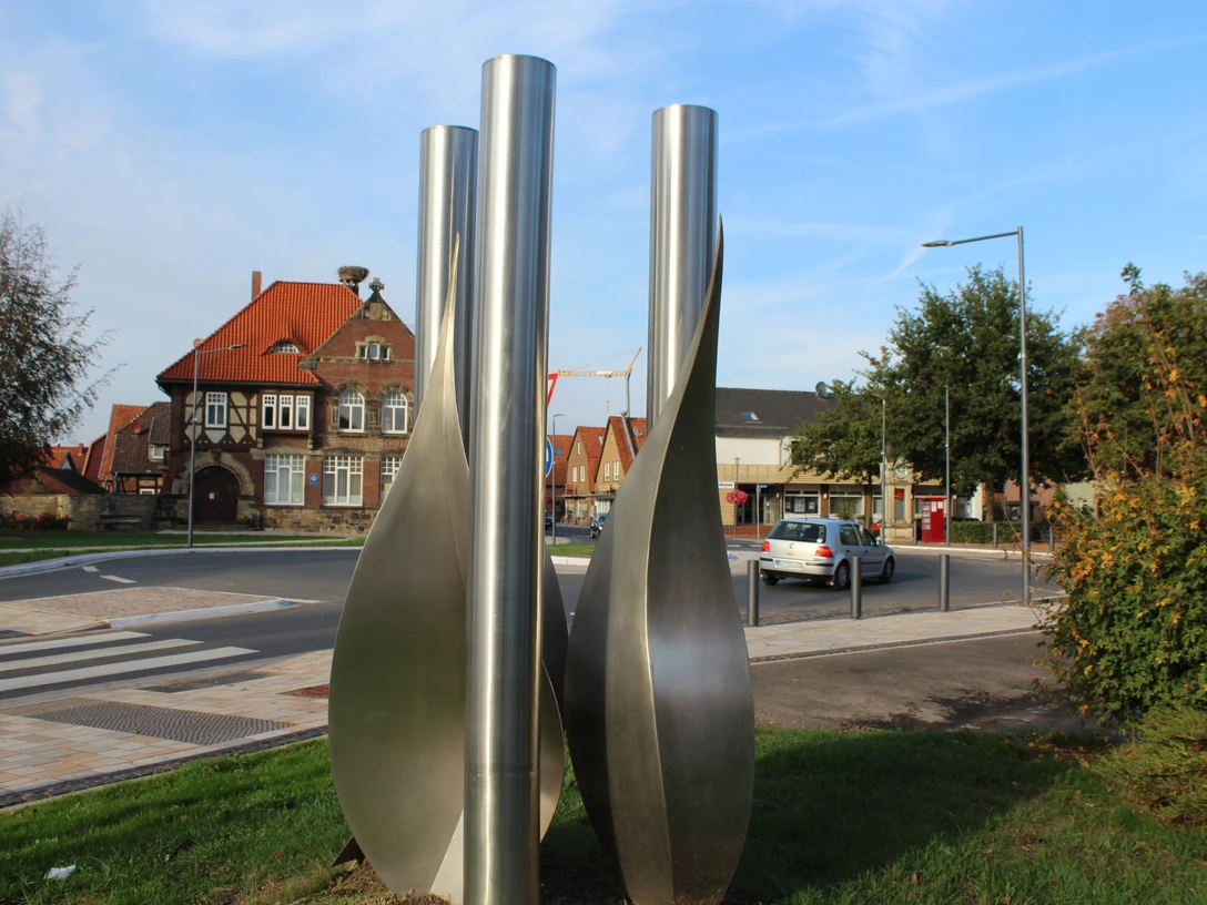 Skulptur Stundenrot schenkt nichts als Blätter Drei hohe, spiralförmige Metallsäulen stehen in einem städtischen Umfeld mit traditionellen Gebäuden.