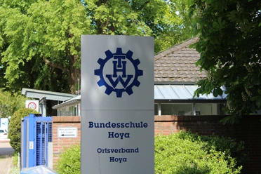 Schild der THW Bundesschule Hoya mit Logo, vor einem Ziegelgebäude und grüner Vegetation.
