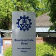 Schild der THW Bundesschule Hoya mit Logo, vor einem Ziegelgebäude und grüner Vegetation.