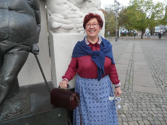 Eine lächelnde Frau in traditioneller Kleidung neben einer Skulptur auf einem gepflasterten Platz.