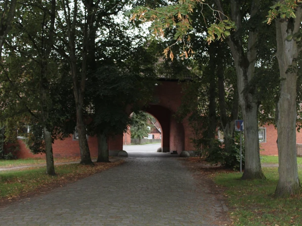 Ein gemauerter Torbogen führt durch eine mit Kieseln gepflasterte Straße, die von Bäumen gesäumt ist.