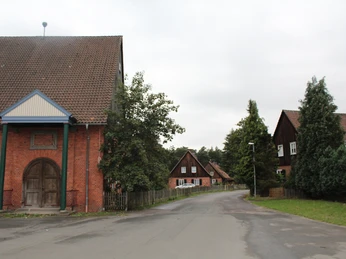 Rote Backsteingebäude mit Satteldächern säumen eine ruhige Straße, umgeben von Bäumen und Wiesen.