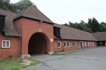 Eingangsbereich eines historischen, roten Backsteingebäudes mit großem Torbogen in einer Waldumgebung.