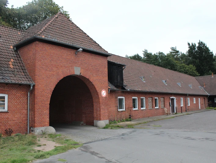 Eingangsbereich eines historischen, roten Backsteingebäudes mit großem Torbogen in einer Waldumgebung.