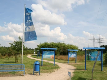 Infotafeln Blauer Pfad mit Infotafeln und Flagge in einer grünen Landschaft unter leicht bewölktem Himmel.