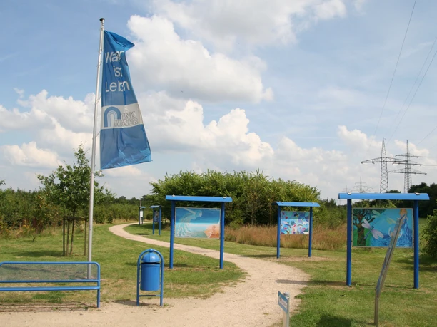 Infotafeln Blauer Pfad mit Infotafeln und Flagge in einer grünen Landschaft unter leicht bewölktem Himmel.