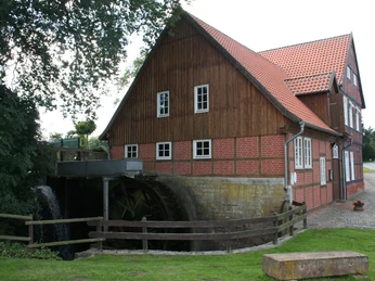 Alte zweigeschossige Wassermühle mit rotem Ziegeldach und Mühlrad, umgeben von Grün und Bäumen.