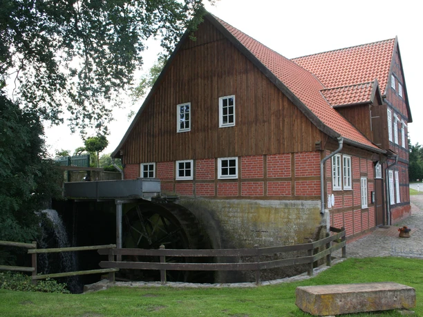 Wassermühle Bruchmühlen Alte zweigeschossige Wassermühle mit rotem Ziegeldach und Mühlrad, umgeben von Grün und Bäumen.