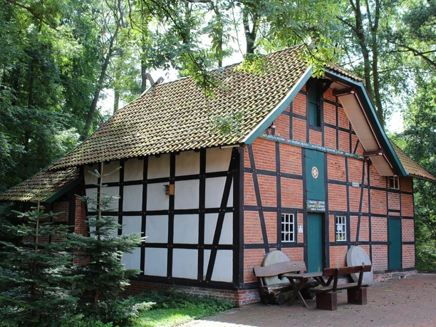The half-timbered building of the Harrienstedt watermill, surrounded by trees, with a wooden bench in front of it.