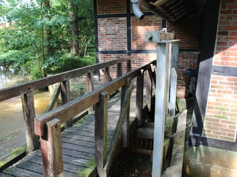 Holzbrücke führt zu Fachwerkbau einer Wassermühle, umgeben von grünem Laub und fließendem Wasser.