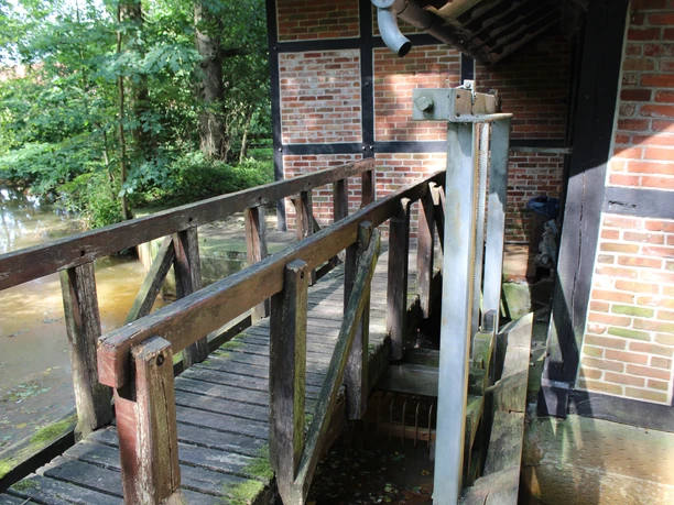 Holzbrücke führt zu Fachwerkbau einer Wassermühle, umgeben von grünem Laub und fließendem Wasser.