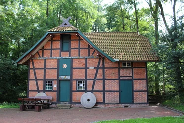 Backsteingebäude mit Fachwerkstrukturen, grünem Dach und zwei Mühlsteinen. Umgebung von Bäumen umgeben.