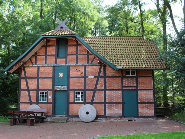 Backsteingebäude mit Fachwerkstrukturen, grünem Dach und zwei Mühlsteinen. Umgebung von Bäumen umgeben.