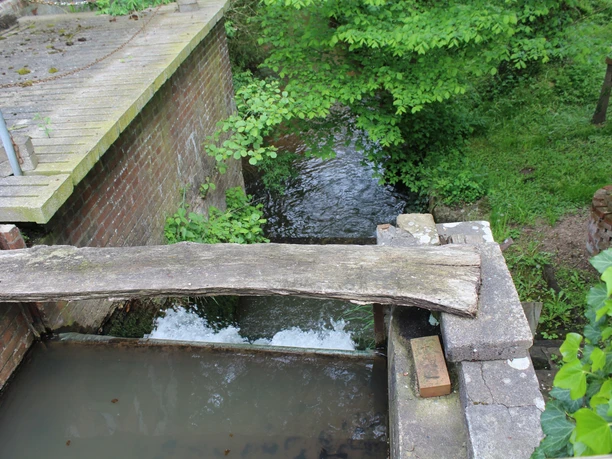 Wassermühle mit hölzerner Brücke, Bach fließt zwischen Ziegelmauern und dichtem, grünem Blattwerk.