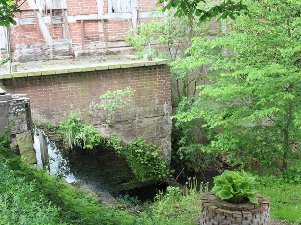 Wassermühle Heuermann Historische Wassermühle mit Backsteingebäude, Wasserfall an grün bewachsenem Mühlrad, umgeben von Bäumen.
