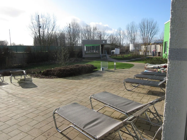 Ontspanningsruimte met verschillende ligstoelen, bomen en een klein saunahuisje op de achtergrond.