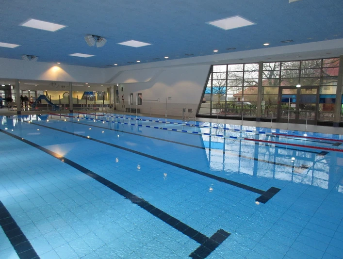 Ein Hallenschwimmbad mit Bahnen, hohen Decken und großen Fenstern, das eine helle Atmosphäre bietet.An indoor swimming pool with lanes, high ceilings and large windows, offering a bright atmosphere.En indendørs swimmingpool med baner, højt til loftet og store vinduer, som giver en lys atmosfære.Een overdekt zwembad met banen, hoge plafonds en grote ramen, die een lichte sfeer bieden.