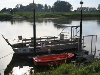 Weserfähre mit rotem Rettungsboot, ruht an einem ruhigen Flussufer, umgeben von grüner Landschaft.