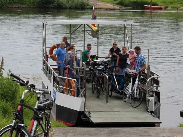 Passagiere mit Fahrrädern stehen auf einer Weserfähre vor einer grünen Flusslandschaft.