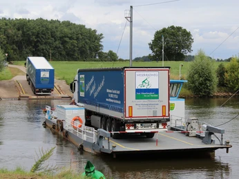 Ein LKW befindet sich auf einer kleinen Fähre, die eine ruhige Flussüberquerung in ländlicher Umgebung vollzieht.