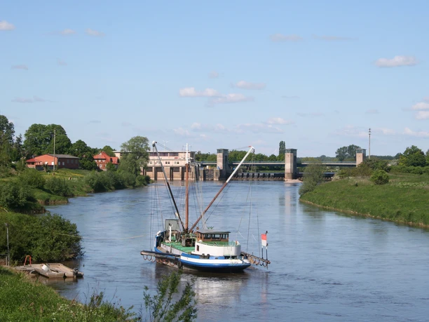 Ein traditionelles Fischerboot, der Aalschocker, liegt vor einem Wehr bei sonnigem Wetter auf einem Fluss.