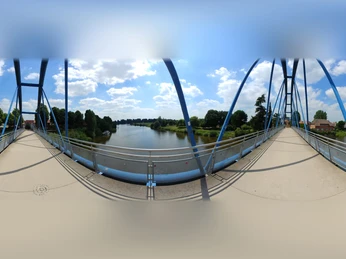 Blick von der Wesertorbrücke in Minden auf die Weser, flankiert von grünen Bäumen und blauem Himmel.