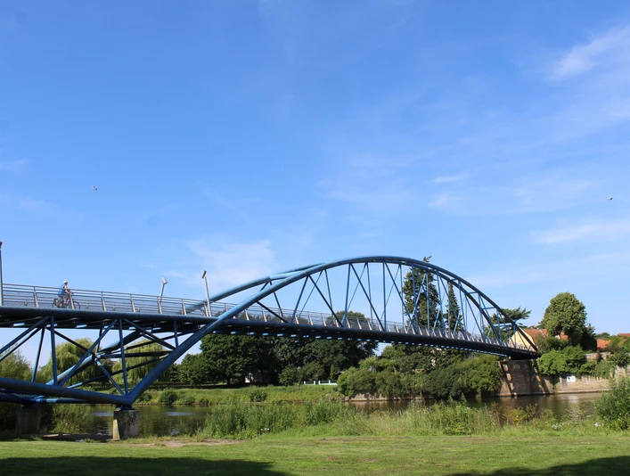 Eine geschwungene blaue Fußgängerbrücke überspannt einen Fluss, umgeben von grüner Vegetation.