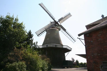 Windmühle Dörverden Windmühle mit weiß-roten Flügeln in ländlicher Umgebung, umgeben von Bäumen und einem Backsteingebäude.