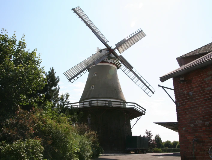 Windmühle Dörverden Windmühle mit weiß-roten Flügeln in ländlicher Umgebung, umgeben von Bäumen und einem Backsteingebäude.