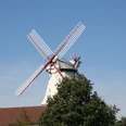 Windmühle "Jan Wind" in Langwedel-Etelsen Windmühle mit roten Flügeln und weißen Holzplanken hinter Bäumen vor blauem Himmel in Langwedel-Etelsen.