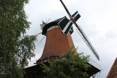 Backstein-Windmühle mit weiß-grünen Flügeln, umgeben von grünen Bäumen, unter bewölktem Himmel.