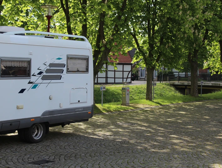 Wohnmobil steht auf einem gepflasterten Platz, umgeben von grünen Bäumen und traditioneller Architektur.