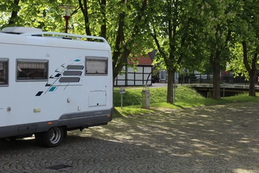 Reisemobillstellplatz Uchte Wohnmobil steht auf einem gepflasterten Platz, umgeben von grünen Bäumen und traditioneller Architektur.