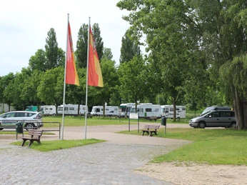 Wohnmobilstellplatz Stolzenau Wohnmobilstellplatz umgeben von grünen Bäumen, mit mehreren geparkten Wohnmobilen und zwei Fahnenmasten im Vordergrund.