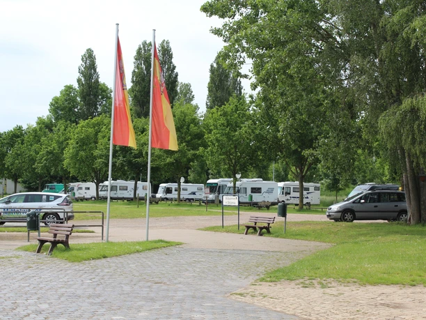 Wohnmobilstellplatz Stolzenau Wohnmobilstellplatz umgeben von grünen Bäumen, mit mehreren geparkten Wohnmobilen und zwei Fahnenmasten im Vordergrund.