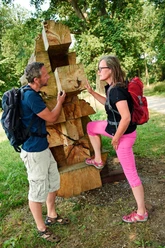 Wanderer am Kunstobjekt "Reise und Heimkehr" Zwei Wanderer betrachten interessiert ein massives Kunstwerk aus Holzscheiten im Freien.