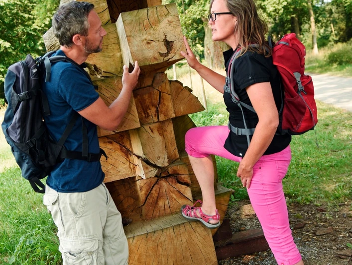 Wanderer am Kunstobjekt "Reise und Heimkehr" Zwei Wanderer betrachten interessiert ein massives Kunstwerk aus Holzscheiten im Freien.