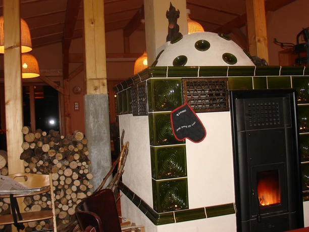 Een gezellige kamer met een tegelkachel, groen geglazuurde tegels, een brandend vuur en een zwarte ovenwant; stapels hout op de achtergrond.