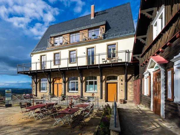 Dvoupatrová budova s balkonem, dřevěnými stoly a židlemi na terase, vedle tradičního dřevěného domu, pod modrou oblohou.