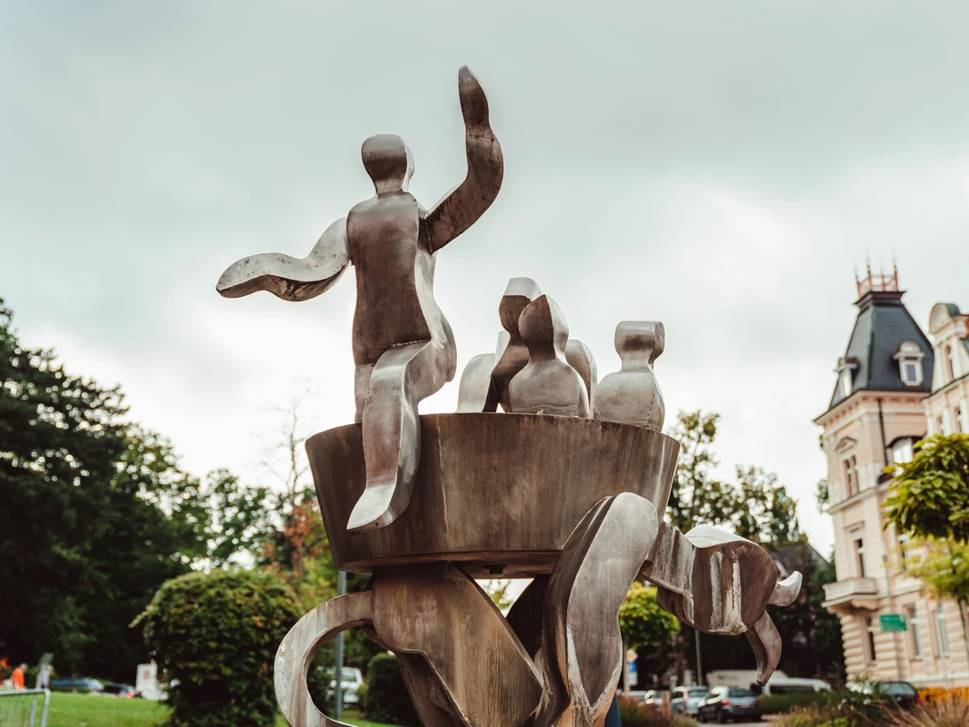 Metallskulptur mit abstrakten Figuren, umgeben von Bäumen und Gebäuden im Zentrum von Bad Nenndorf.
