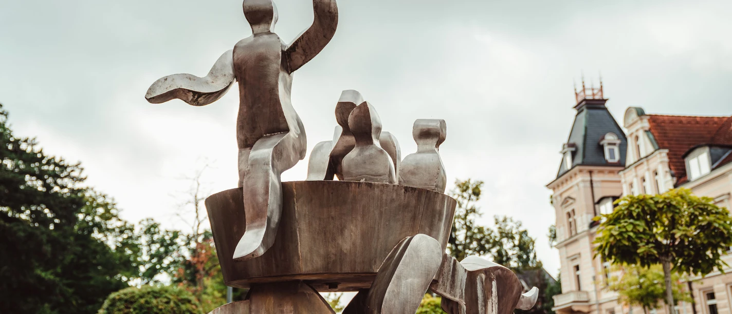 Metallskulptur mit abstrakten Figuren, umgeben von Bäumen und Gebäuden im Zentrum von Bad Nenndorf.