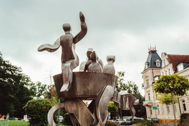 Brunnen "Auf dem Düvelsdreck" Metallskulptur mit abstrakten Figuren, umgeben von Bäumen und Gebäuden im Zentrum von Bad Nenndorf.