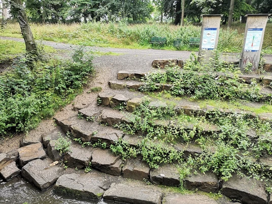 Infotafeln an der Bastau Infotafeln informieren Besucher an einem von Pflanzen gesäumten Wegabschnitt mit Naturstein-Stufen.